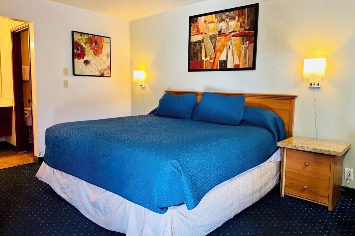 A hotel room with a large bed, a wooden nightstand, two wall-mounted lamps, and two paintings.