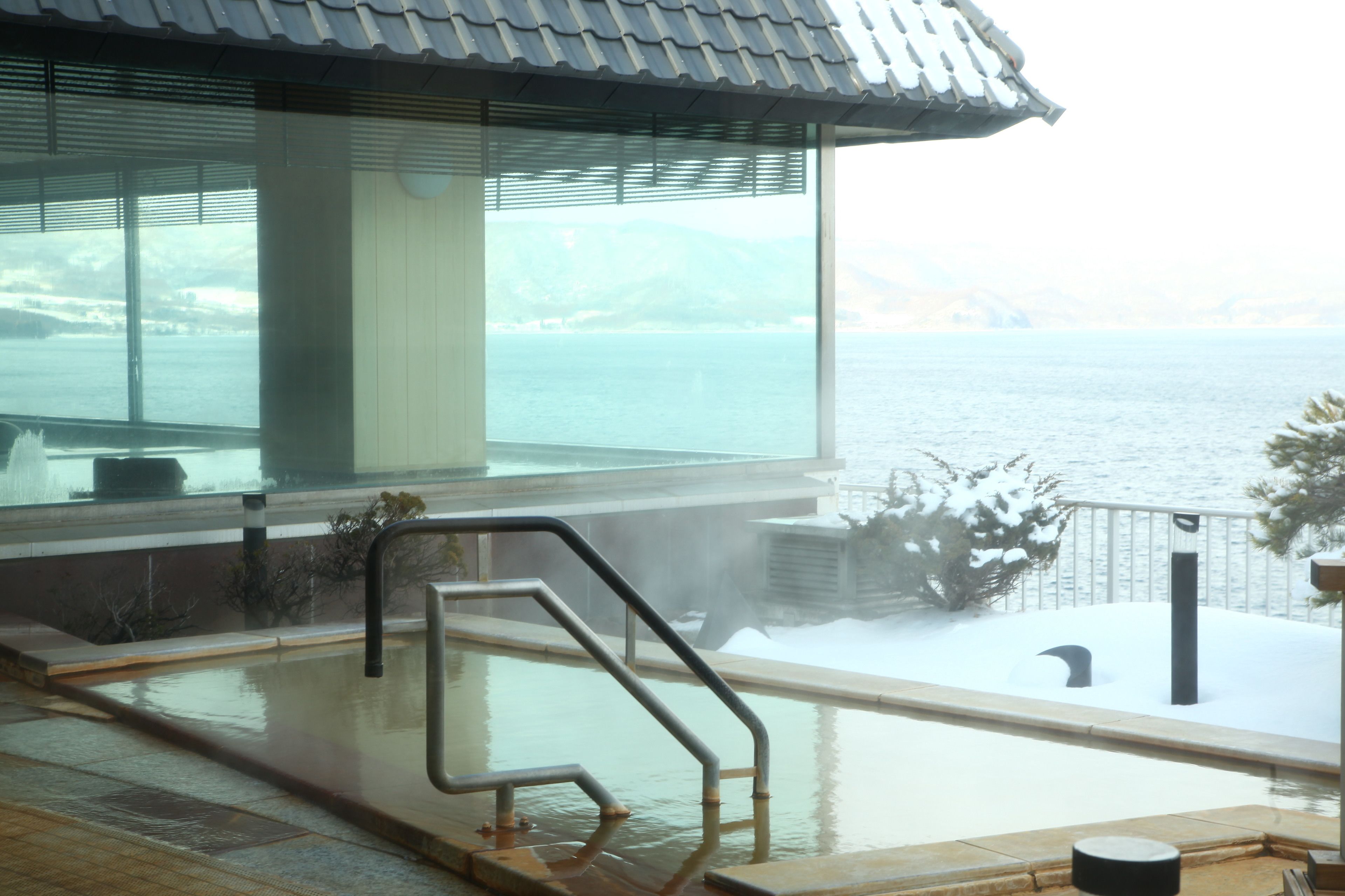 A hot spring area with a view of the sea, a wooden deck, and a traditional tiled roof.