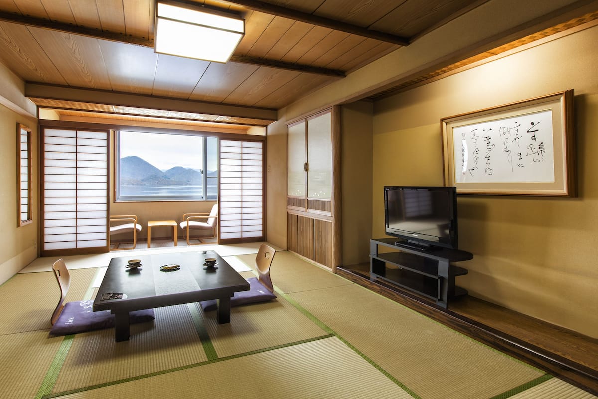 A traditional Japanese room with tatami flooring, a low table, chairs, a television, and a view of mountains through a shoji screen.