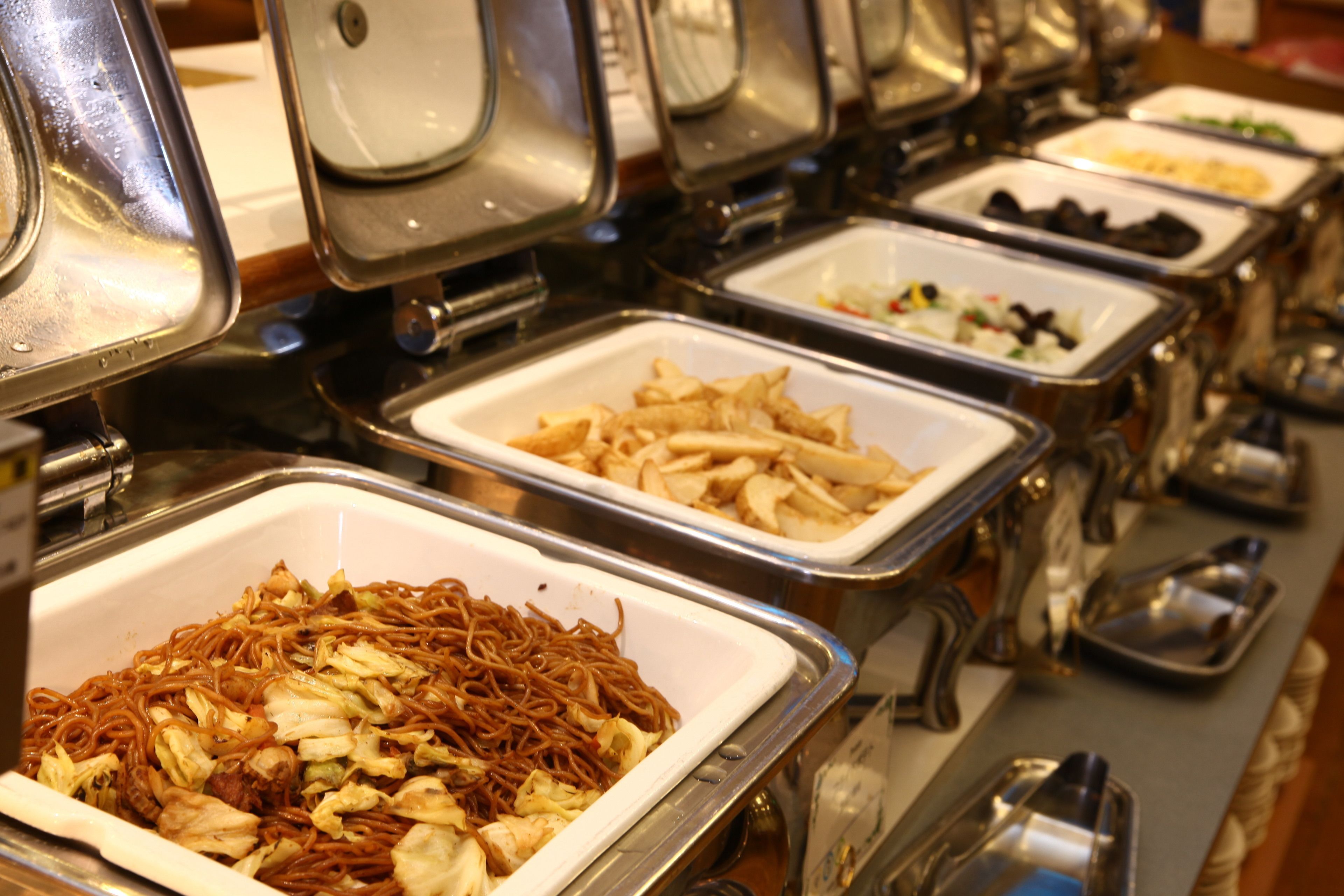Un bufé con diversas opciones de comida, incluyendo fideos, papas fritas y otros platos, en bandejas de metal.