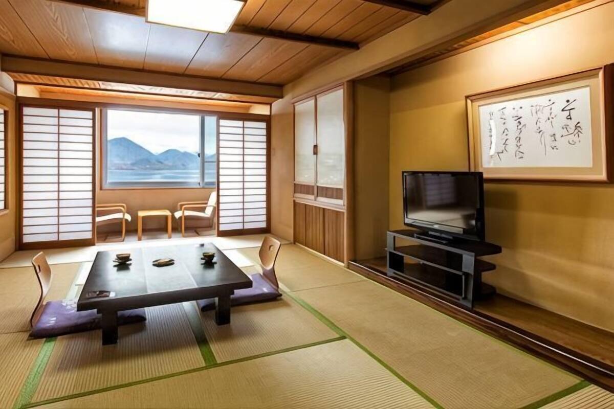 A traditional Japanese room with tatami flooring, a low table, chairs, a TV, and a view of mountains through shoji screens.