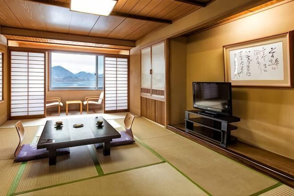 A traditional Japanese room with tatami flooring, a low table, chairs, a TV, and a view of mountains through shoji screens.