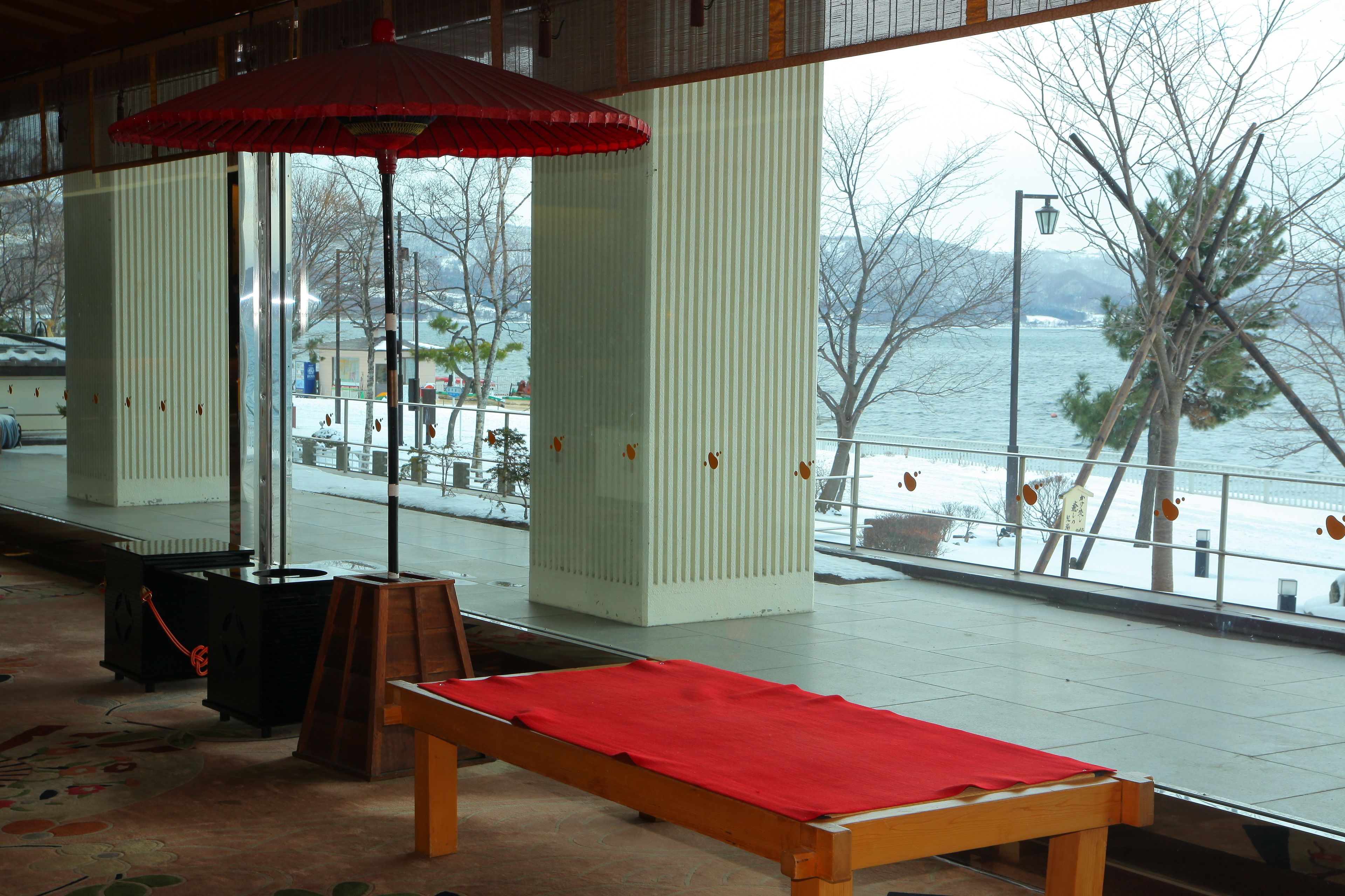 A room with a view of a body of water, a red umbrella, and a wooden bench with a red cloth.