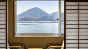 Habitación con ventana grande que ofrece vista a un lago y montañas. Hay dos sillas y una pequeña mesa.