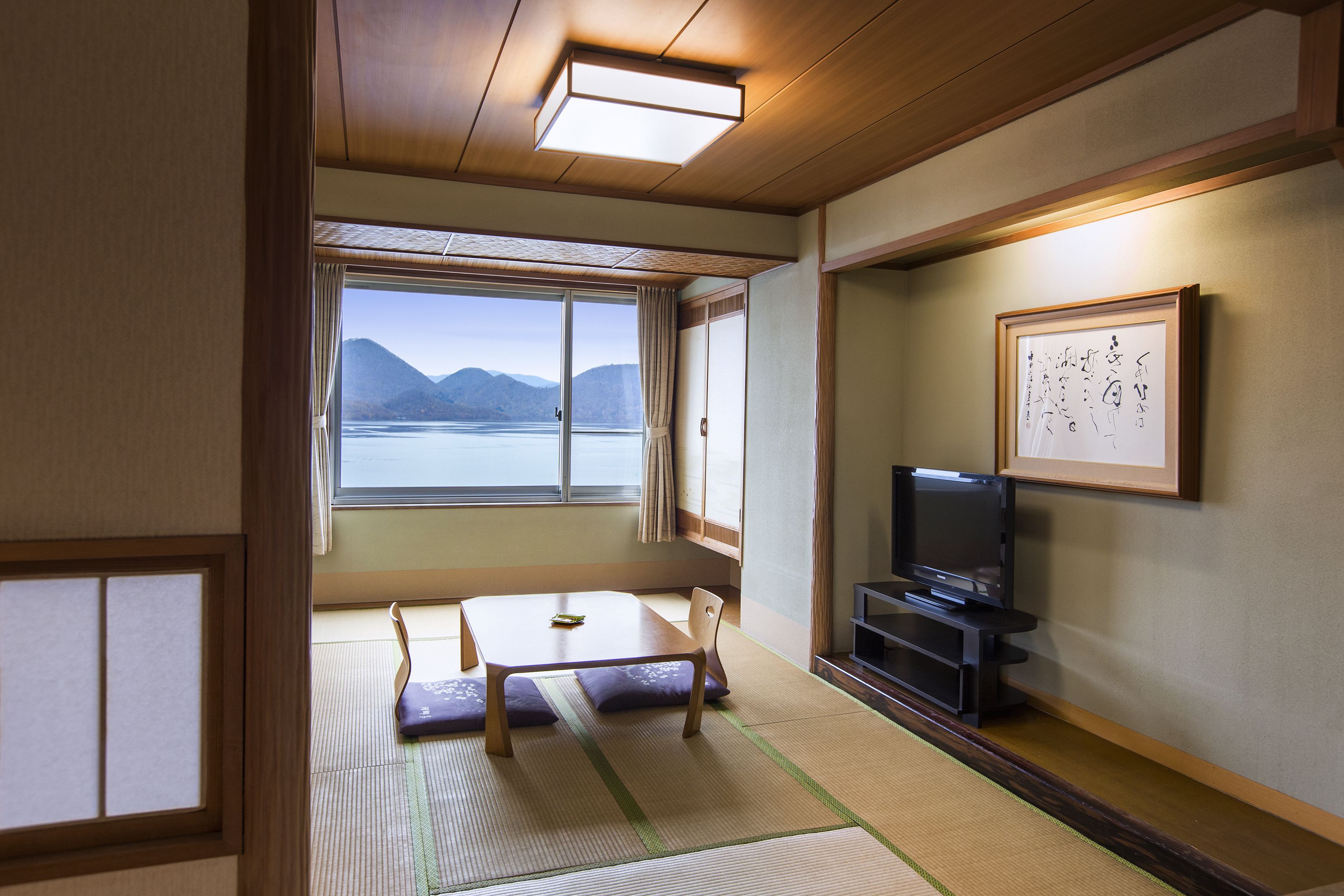 A traditional Japanese room with tatami flooring, a low table, a television, and a view of a lake and mountains.