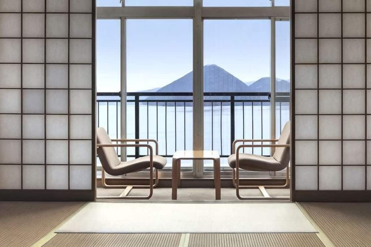 A room with tatami flooring, a sliding shoji screen, two chairs, a table, and a view of a mountain through large windows.