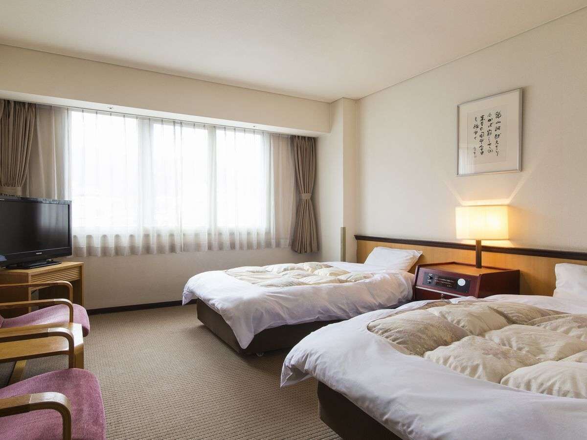Quarto de hotel com duas camas, televisão, mesa de cabeceira com abajur e uma janela grande com cortinas.