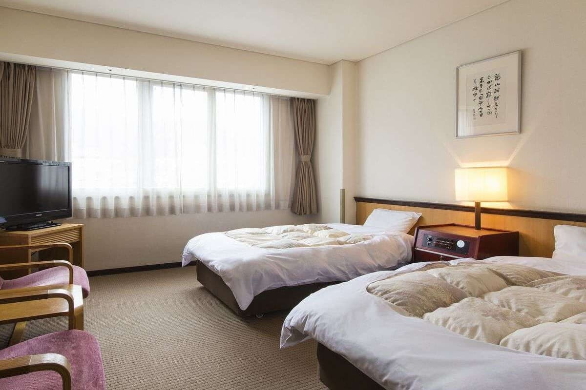 A hotel room with two beds, a TV, a bedside table with a lamp, and a large window with curtains.