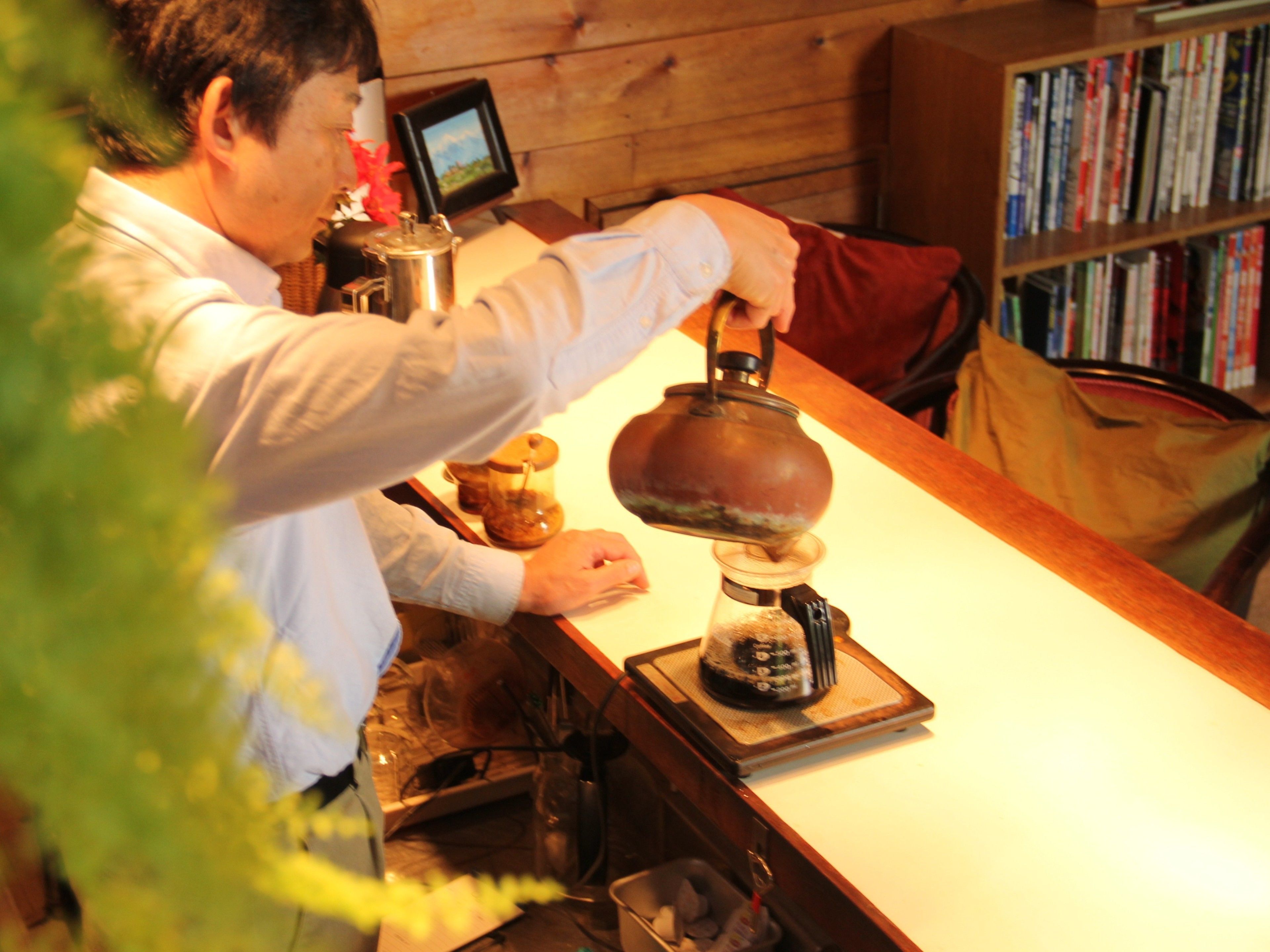 A person at a bar with a kettle, a coffee machine, and a bookshelf in the background.
