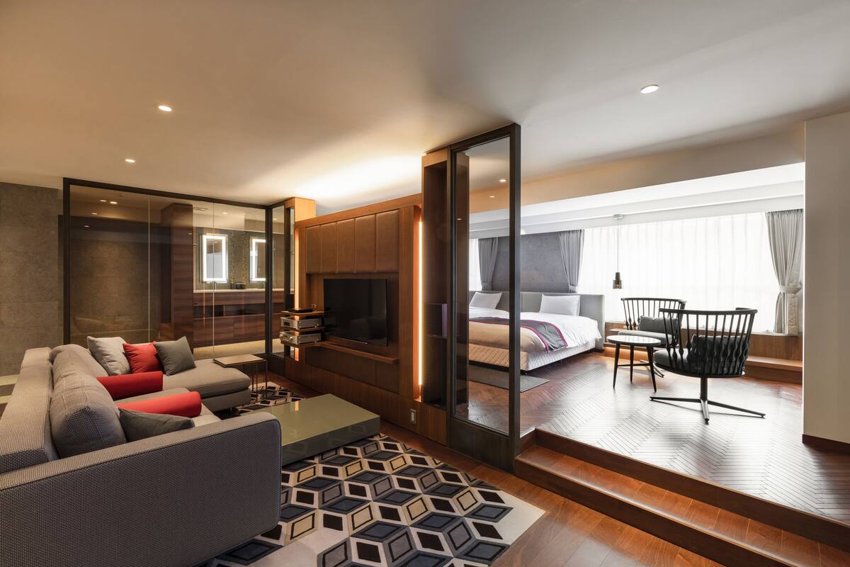 A modern hotel room with a sofa, a TV, a dining area, and a large window.