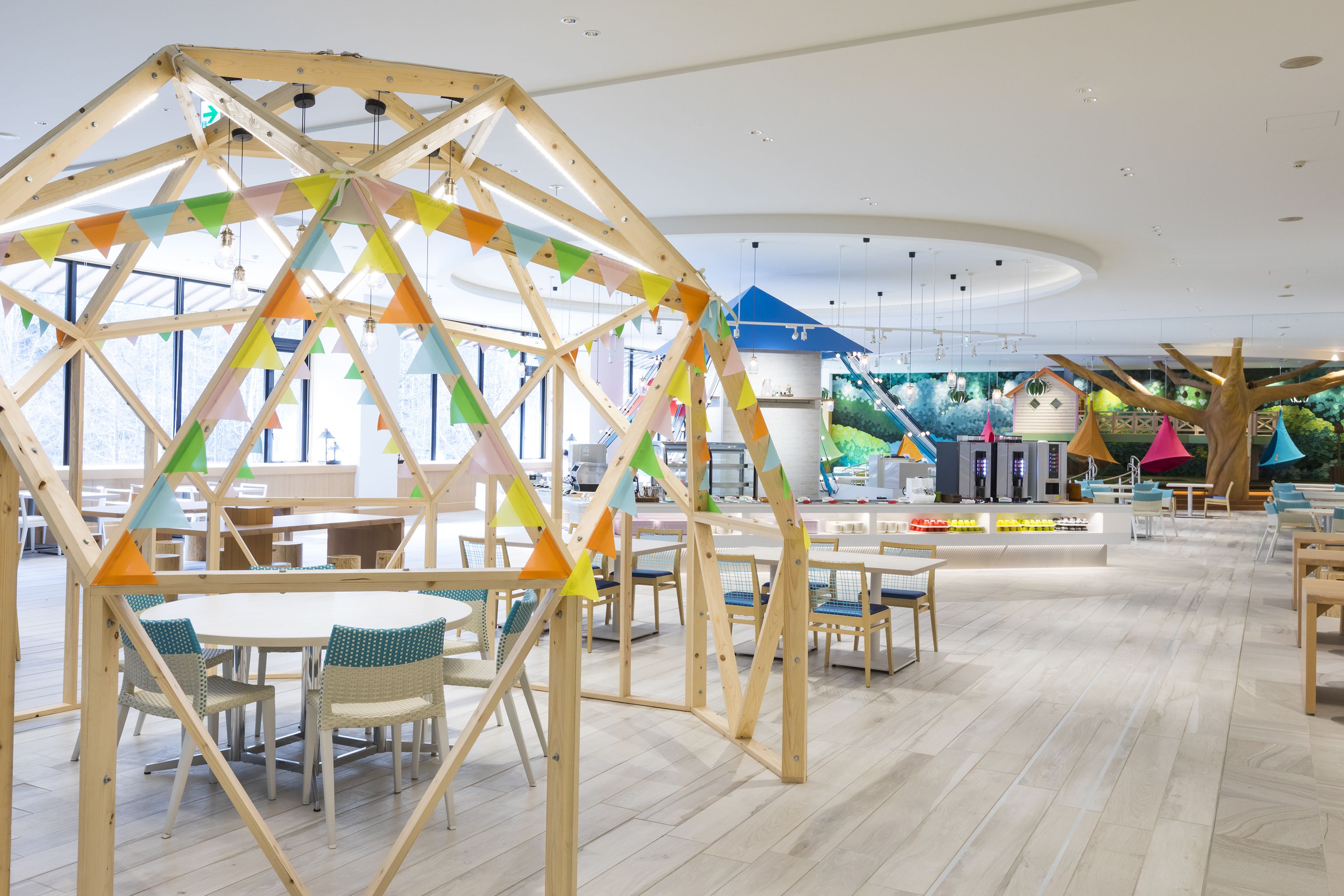 A spacious indoor area with wooden structures and tables, featuring a large window and a play area in the background.