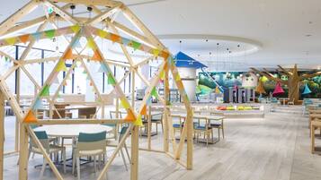 A spacious indoor area with wooden structures and tables, featuring a large window and a play area in the background.