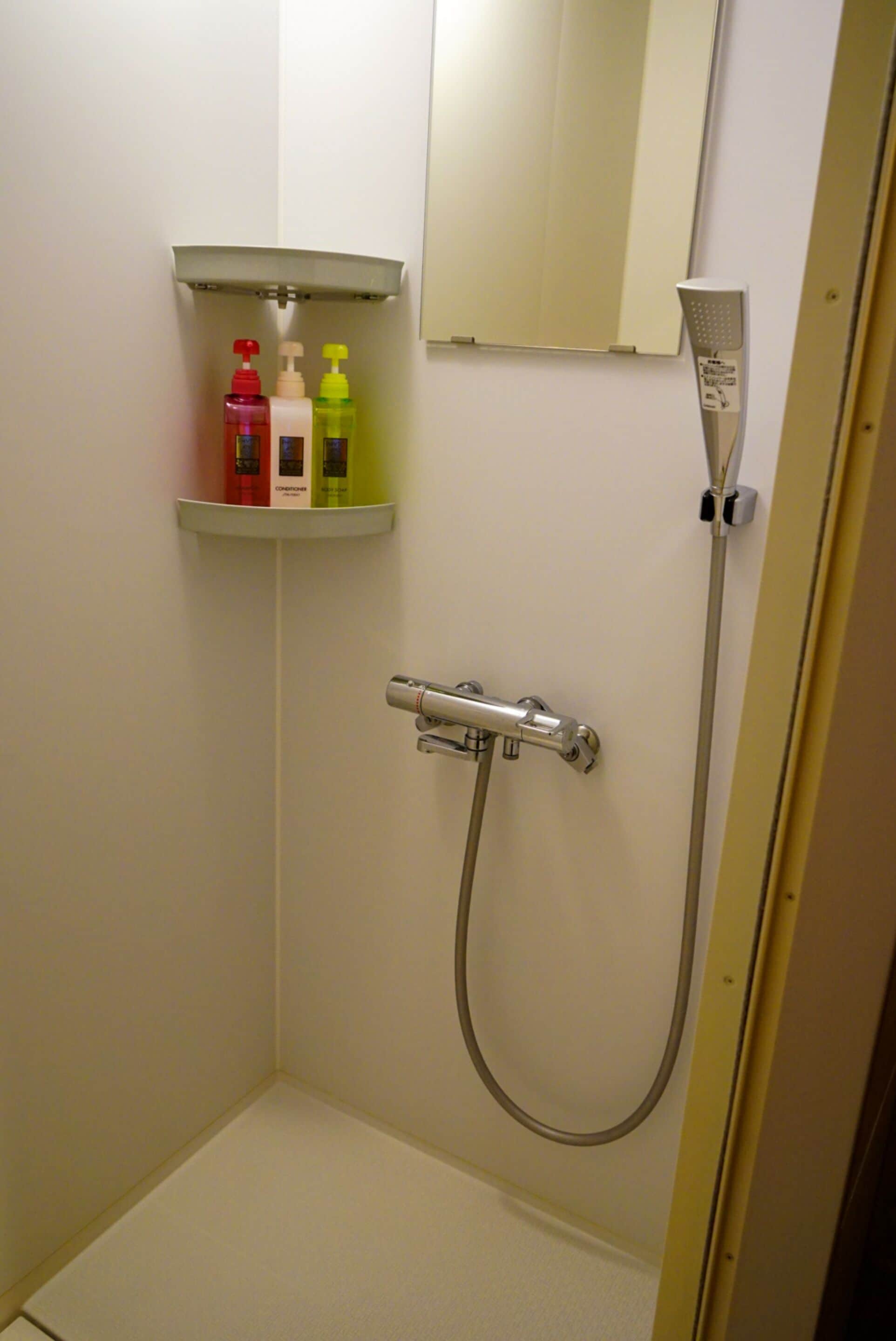 A small bathroom with a shower, a mirror, and a shelf with toiletries.
