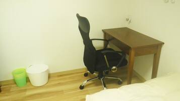 Una habitación pequeña con un escritorio de madera, una silla de oficina negra, un bote de basura verde y blanco, y una cama.