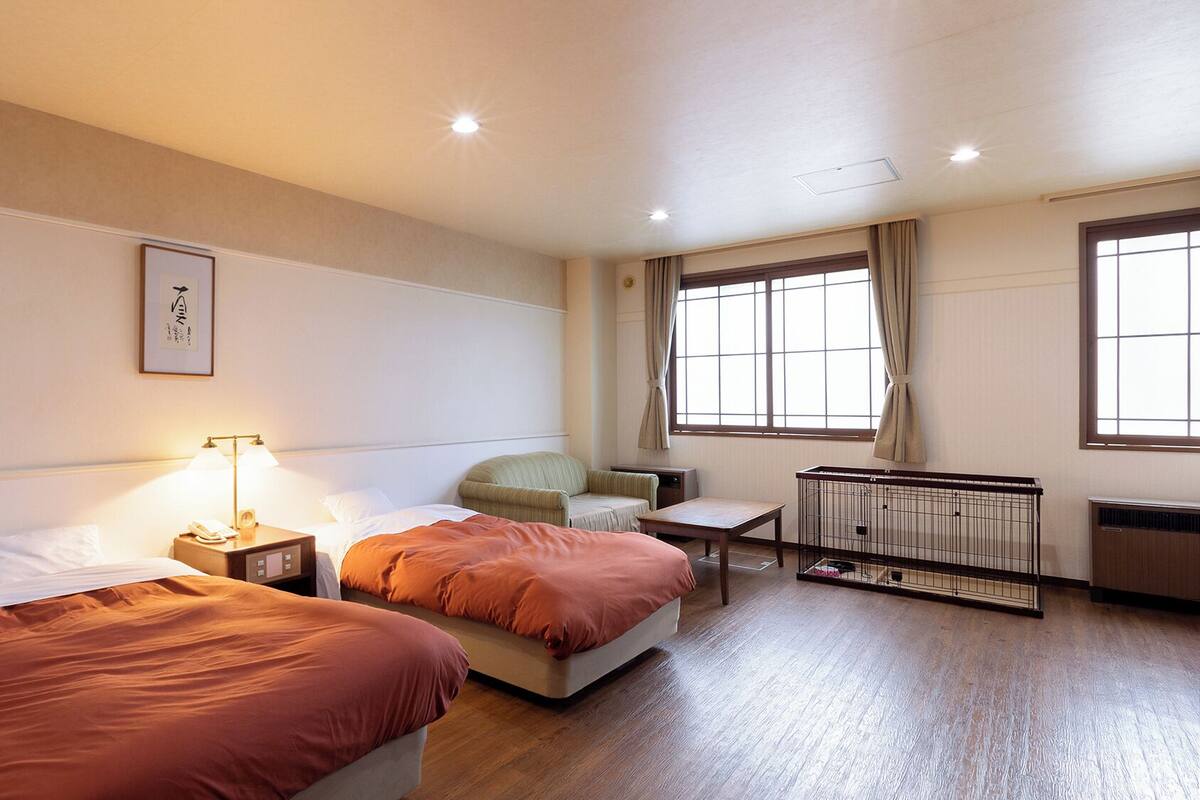 A hotel room with two beds, a small table, a chair, a radiator, and a window with curtains.