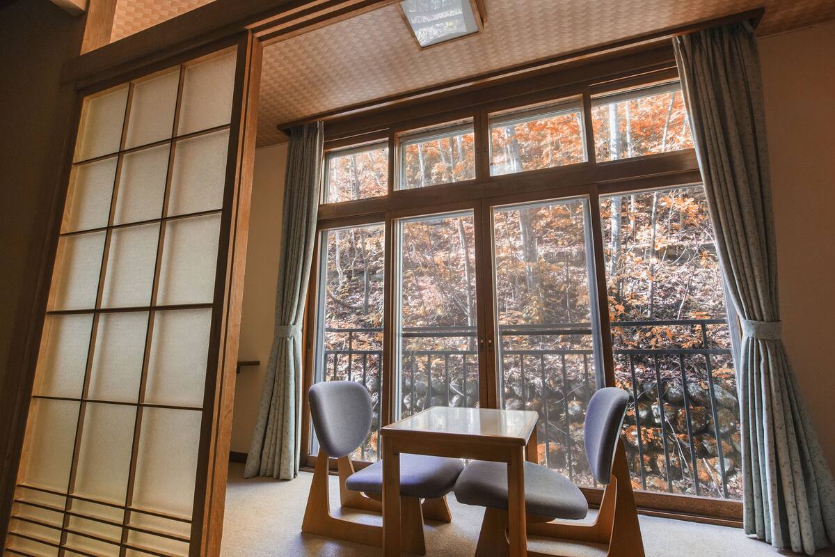 A room with tatami flooring, a low wooden table, two chairs, and a large window with a view of autumn trees.