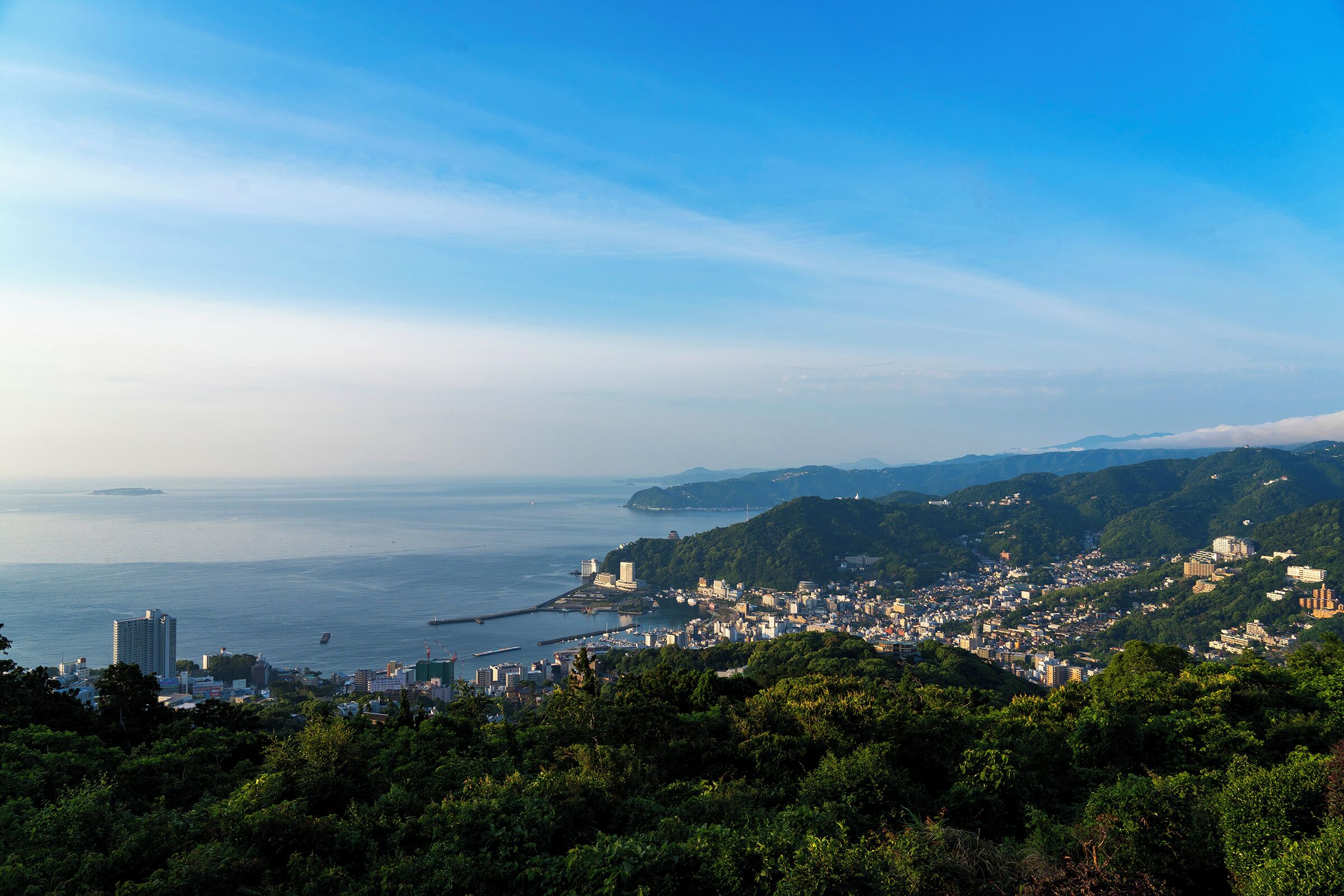 A coastal cityscape with dense urban development, a prominent body of water, and lush green hills in the background.