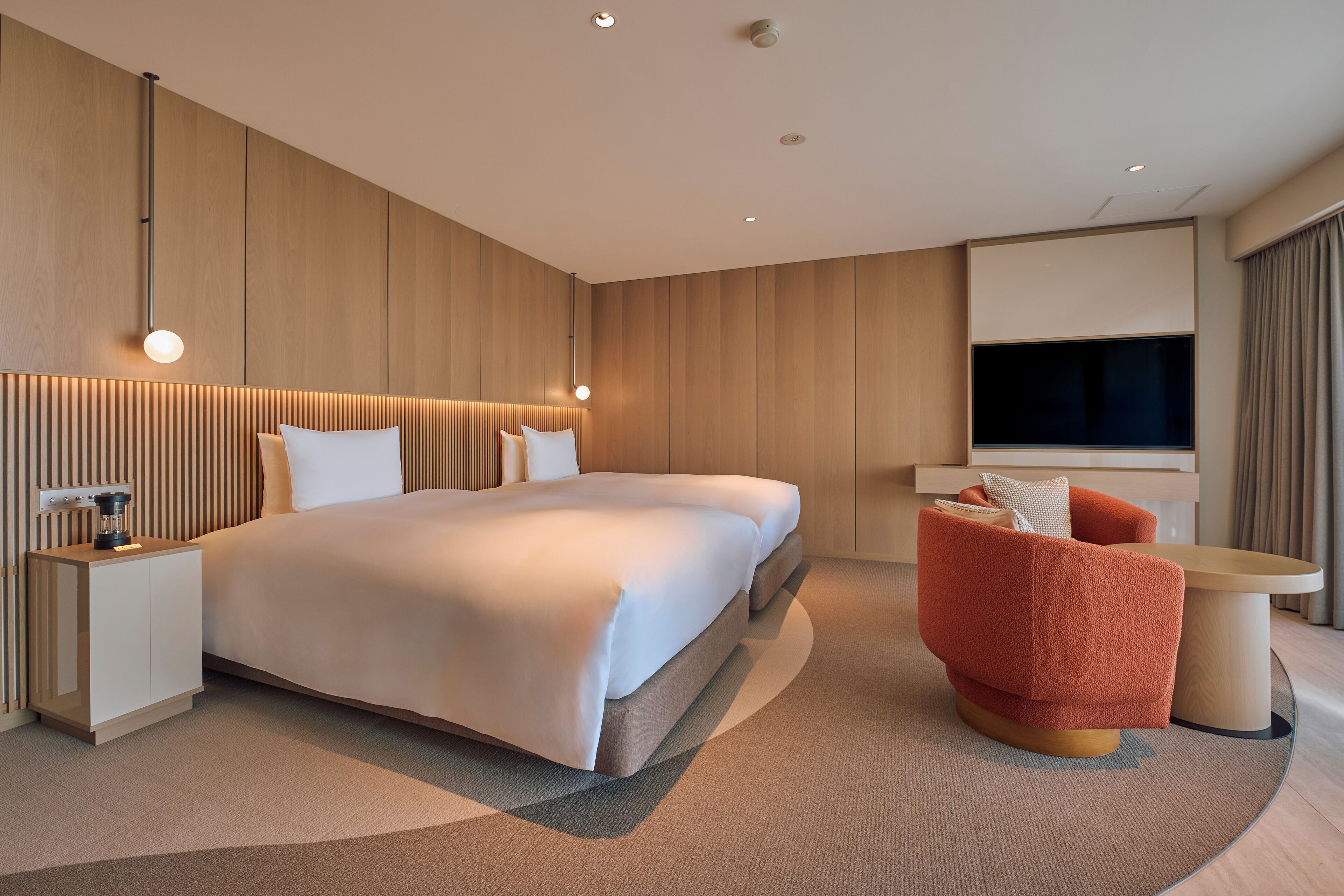 Ein modernes Hotelzimmer mit einem großen Bett, einer Sitzecke mit einem roten Sessel, einem kleinen Tisch und einem an der Wand montierten Fernseher.