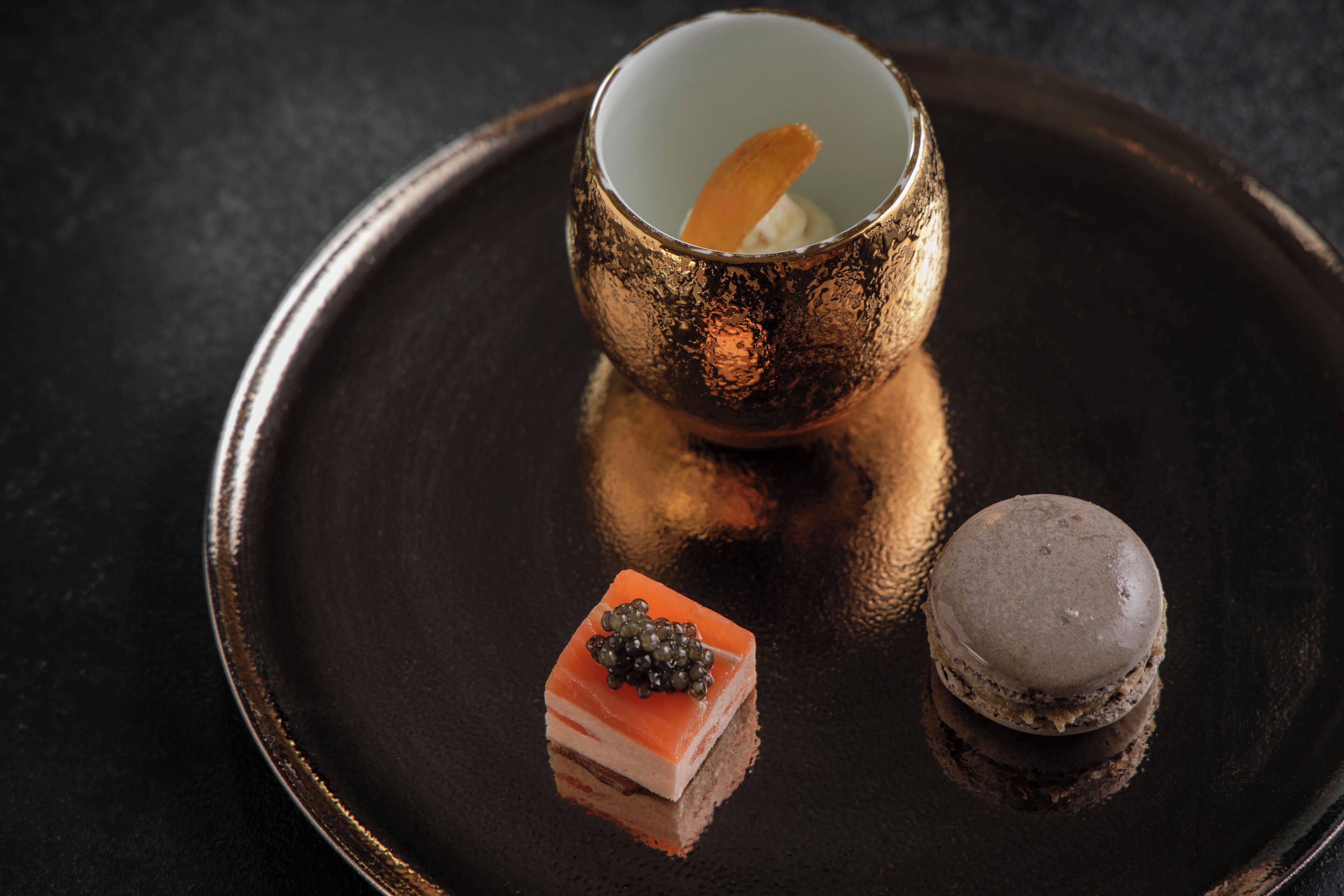 Un plato de postre con un macaron, un trozo de pastel con caviar y una taza con una cuchara.