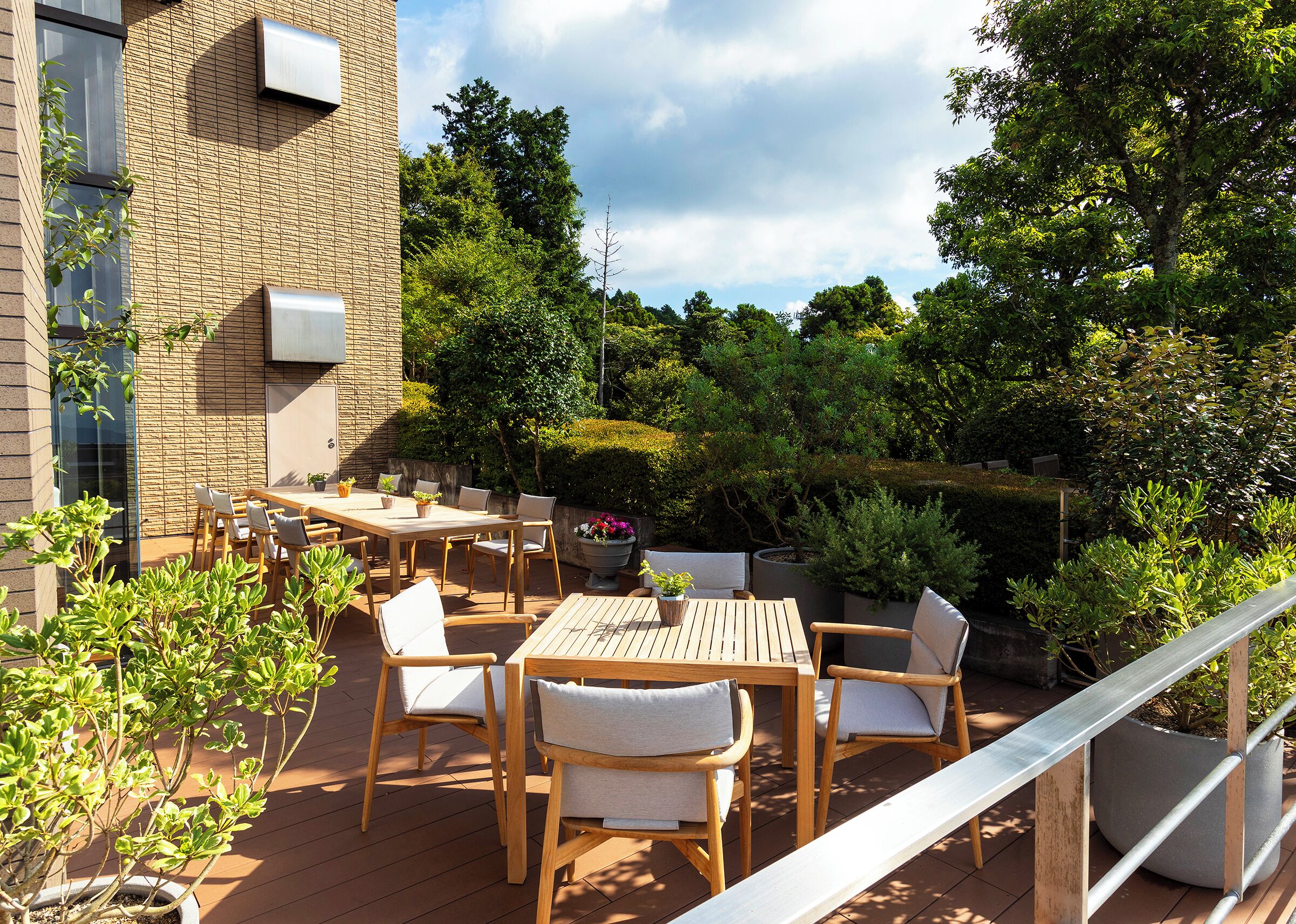 An outdoor patio with wooden tables and chairs, surrounded by potted plants and trees.