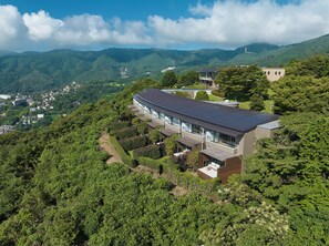 A modern building with a curved roof, surrounded by lush greenery and mountains in the background. - Hotel Grand Bach Atami Crescendo (Atami)