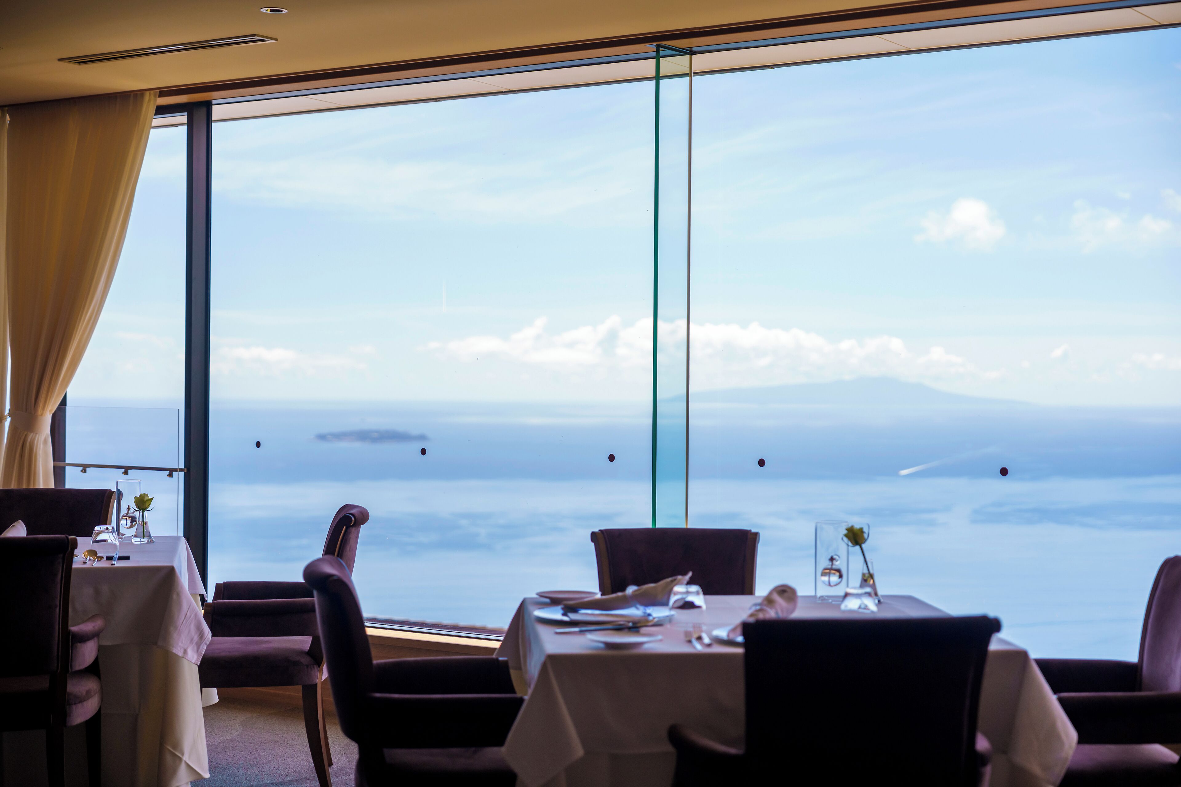 Een eetruimte met een tafel gedekt voor twee personen, grote ramen met uitzicht op zee en verre eilanden.