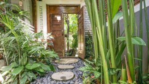 Un sendero de jardín con una puerta de madera, rodeado de abundante follaje y piedras.