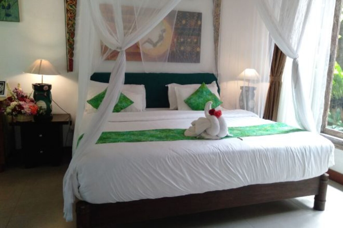 Una cama bien tendida con ropa blanca y detalles verdes, un cojín decorativo con forma de cisne y un dosel.
