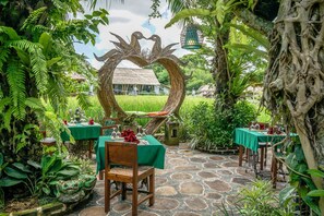 Un área de comedor al aire libre con mesas dispuestas para dos, rodeada de exuberante follaje y un arco de madera con forma de corazón.