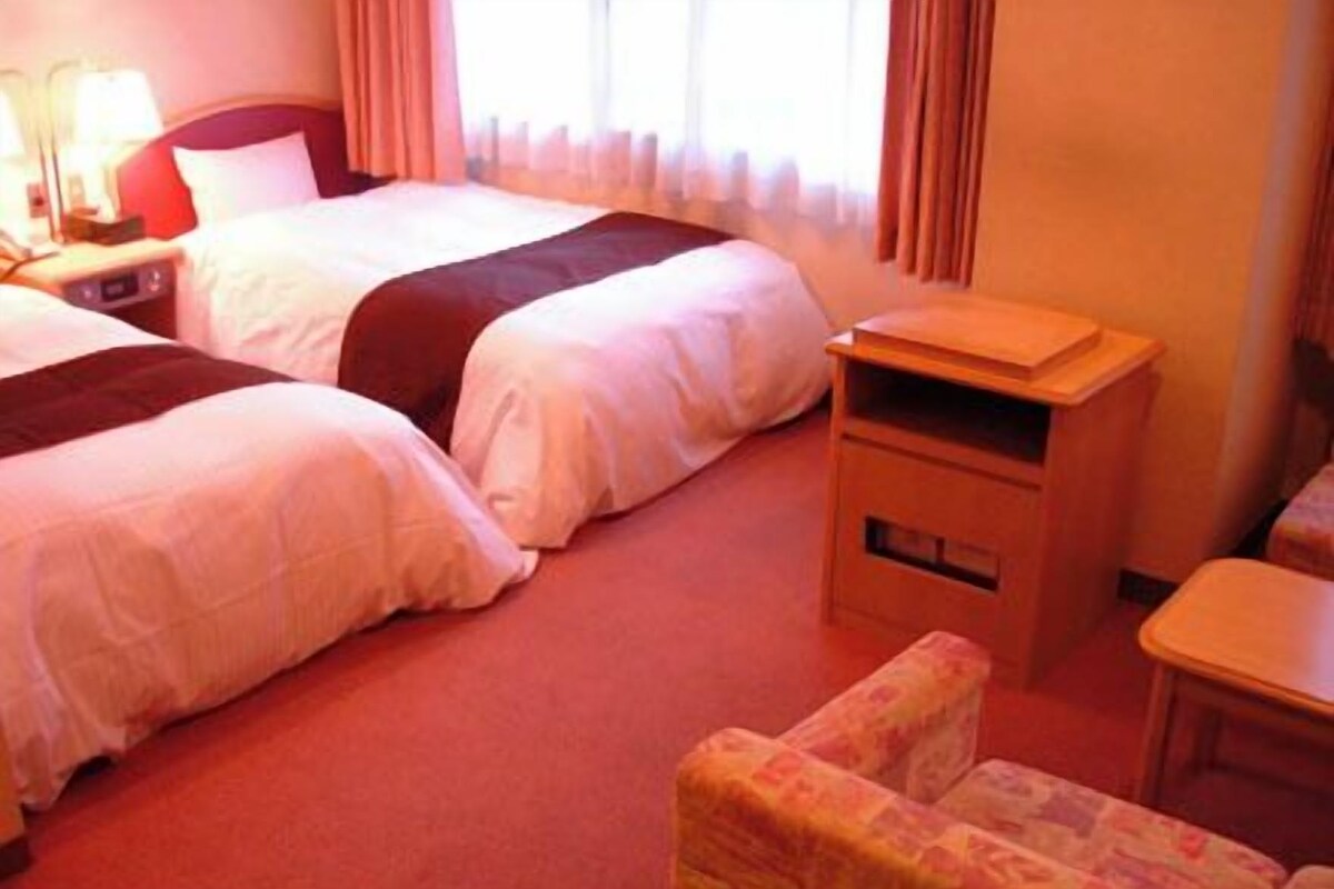 Camera d'albergo con due letti, un comodino con lampada, un tavolino e una sedia.