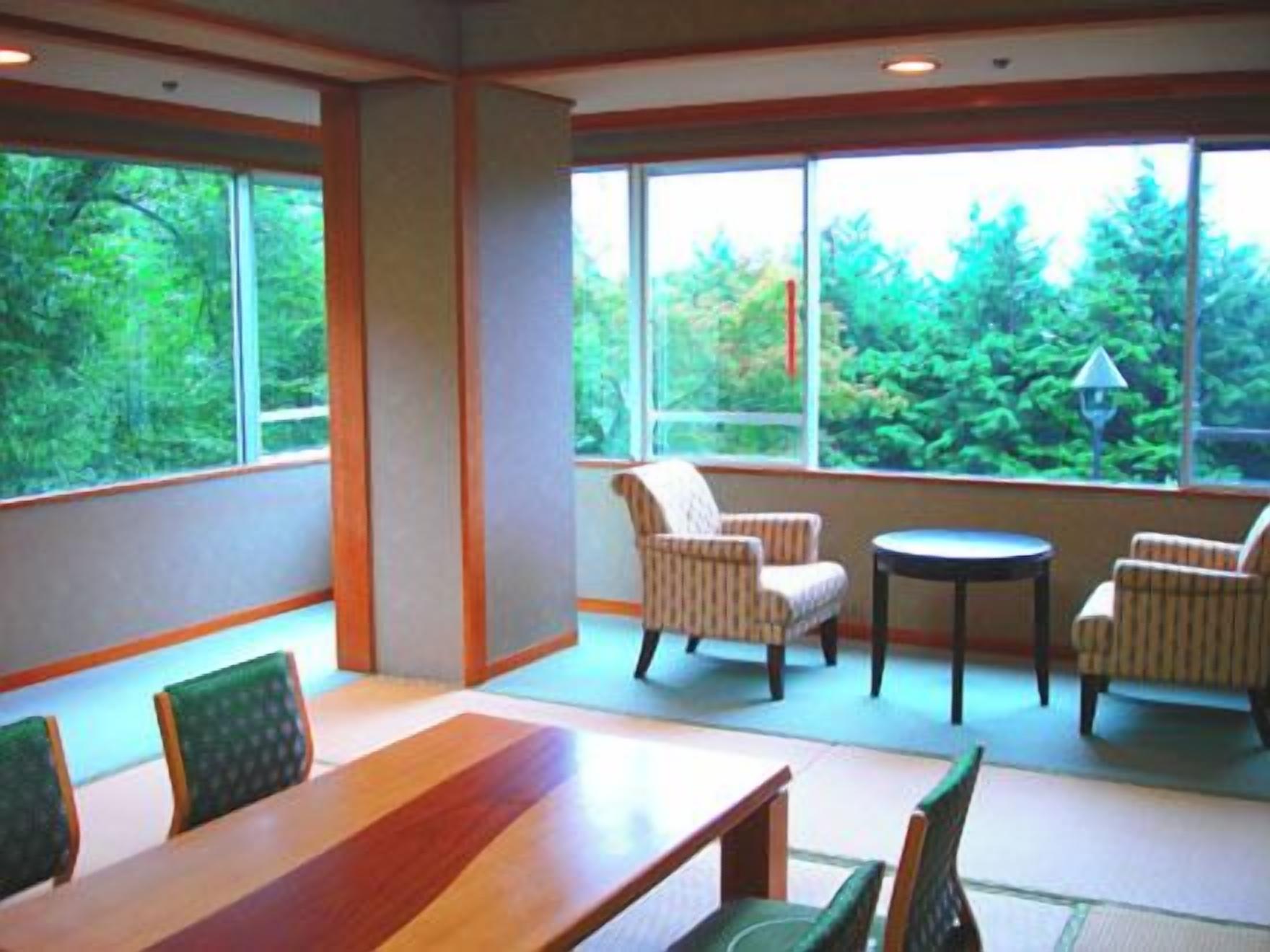 A room with a wooden table and chairs, large windows with a view of greenery, and a small round table with chairs.
