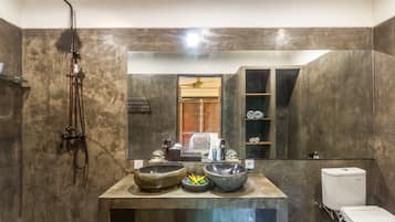 A bathroom with a large mirror, two stone sinks, a showerhead, and a toilet.