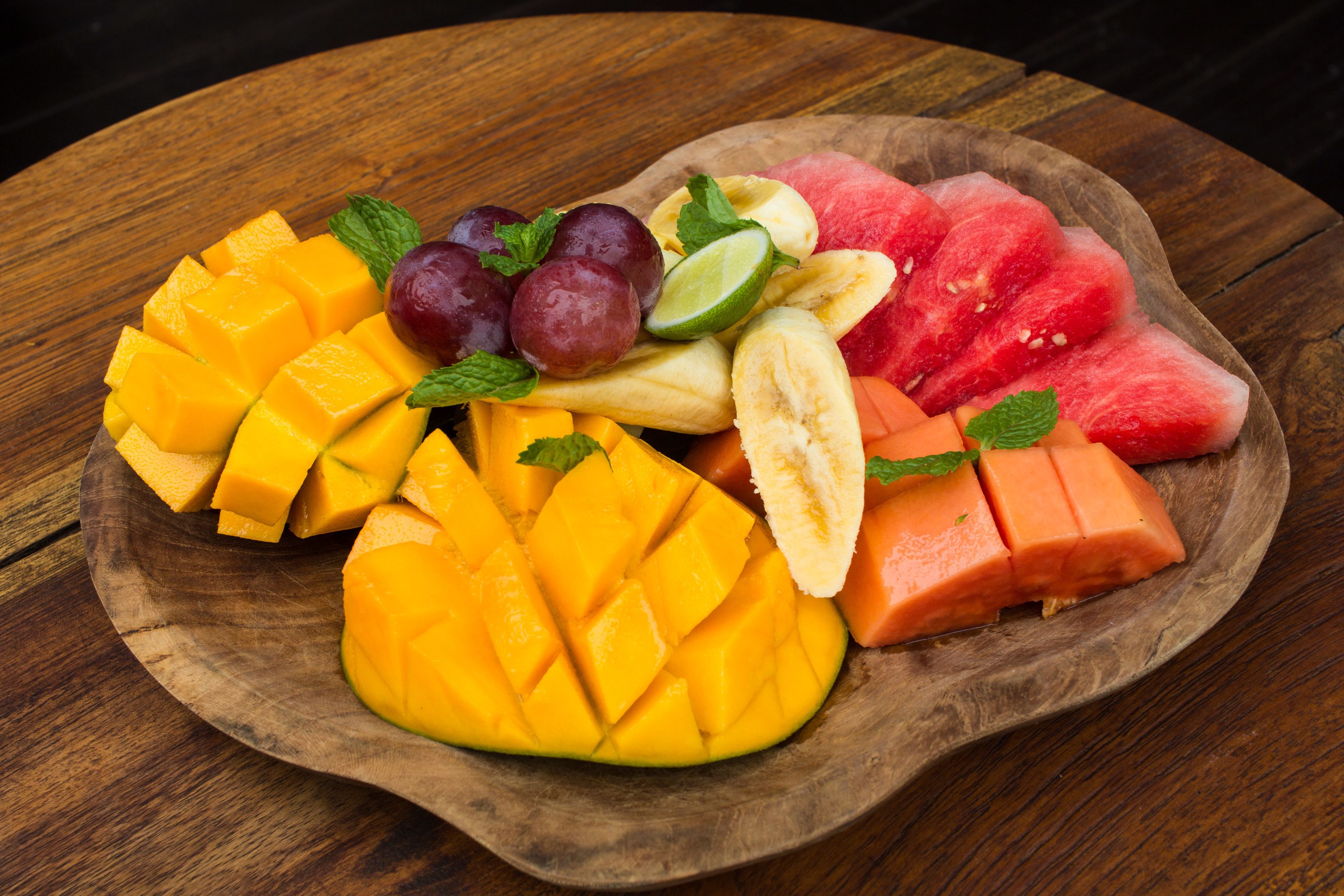 A wooden plate with sliced mango, watermelon, bananas, grapes, and lime on a wooden table.