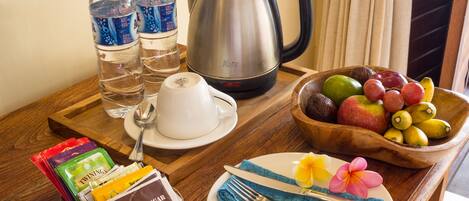 A breakfast setup with a kettle, water bottles, a fruit bowl, a plate with a flower, and packaged snacks.
