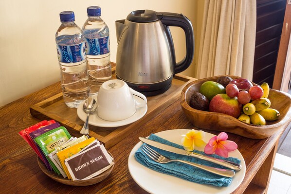 Preparación para el desayuno con una tetera, botellas de agua, un frutero, un plato con una flor y aperitivos empaquetados.