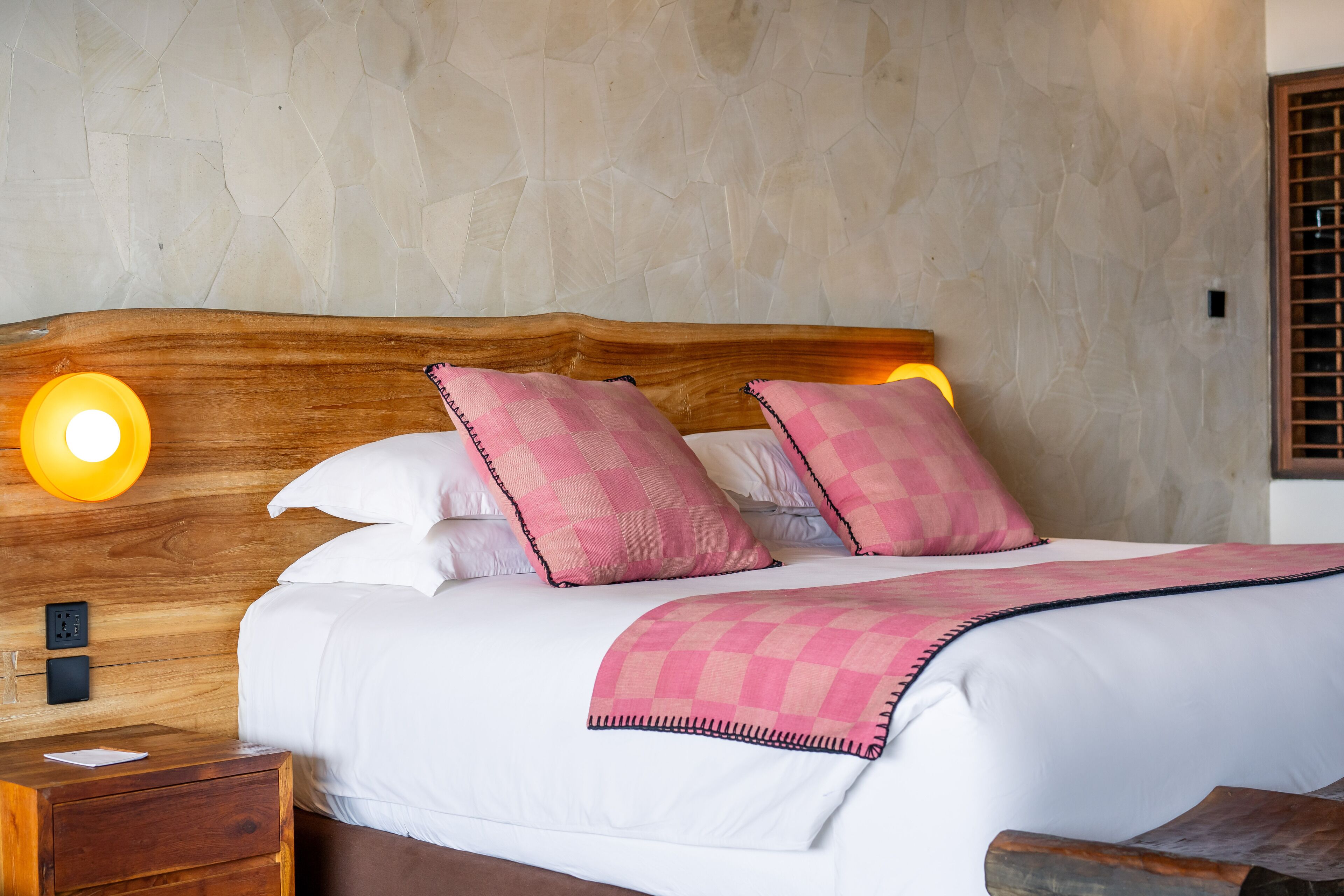A neatly made bed with white linens and pink checkered pillows, a wooden headboard, and two bedside lamps.