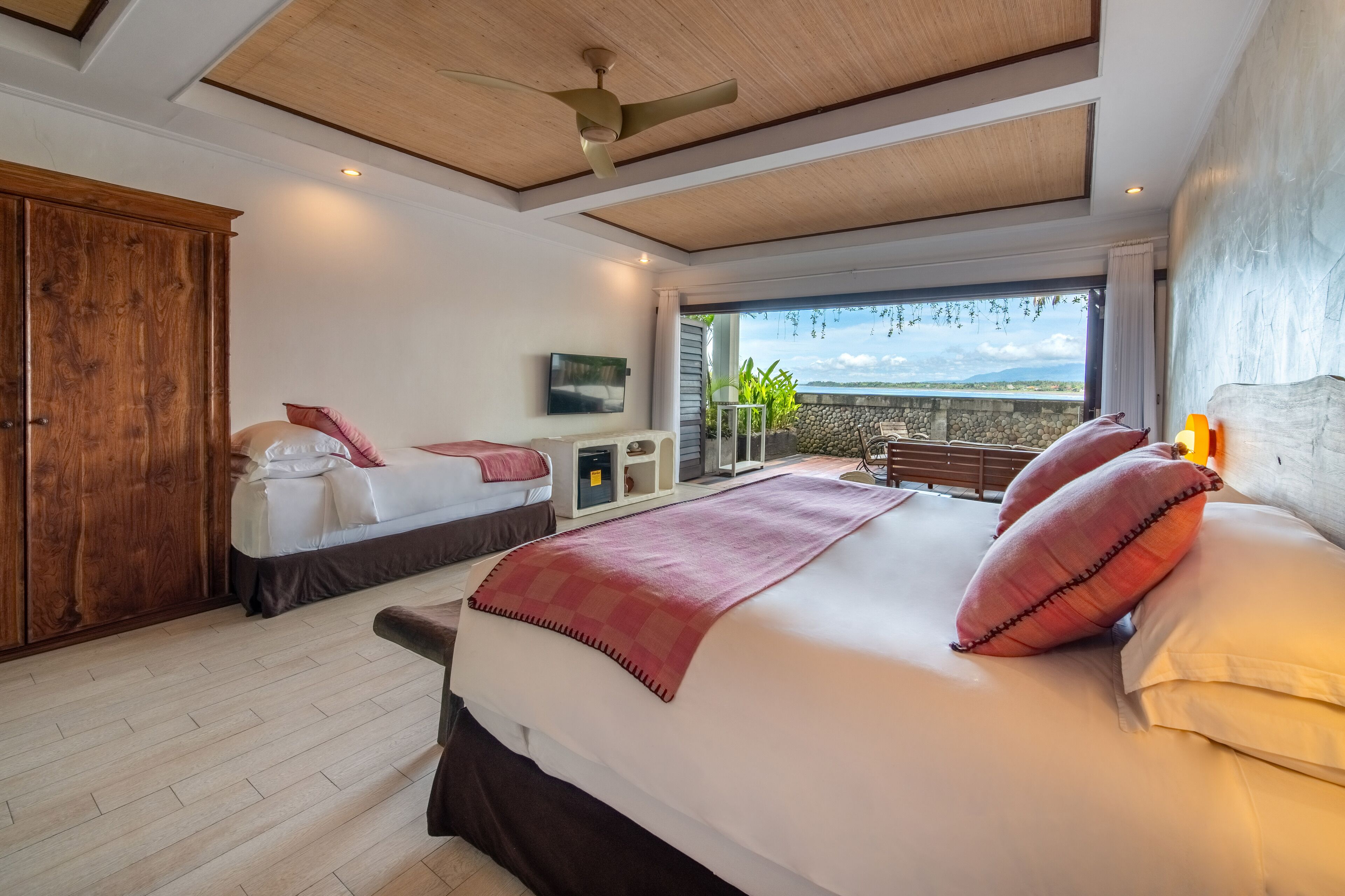 A bedroom with a large bed, wooden wardrobe, ceiling fan, and a view of the outdoors.