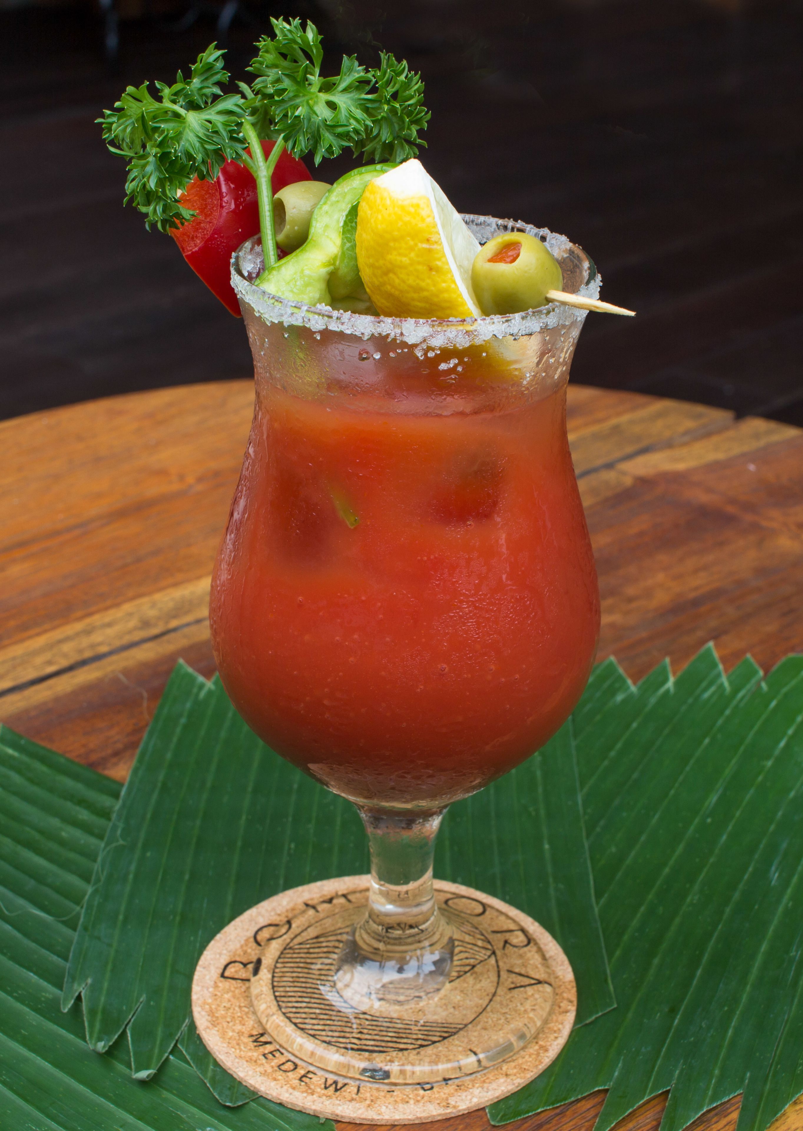 A Bloody Mary cocktail in a stemmed glass with garnishes, served on a wooden coaster.
