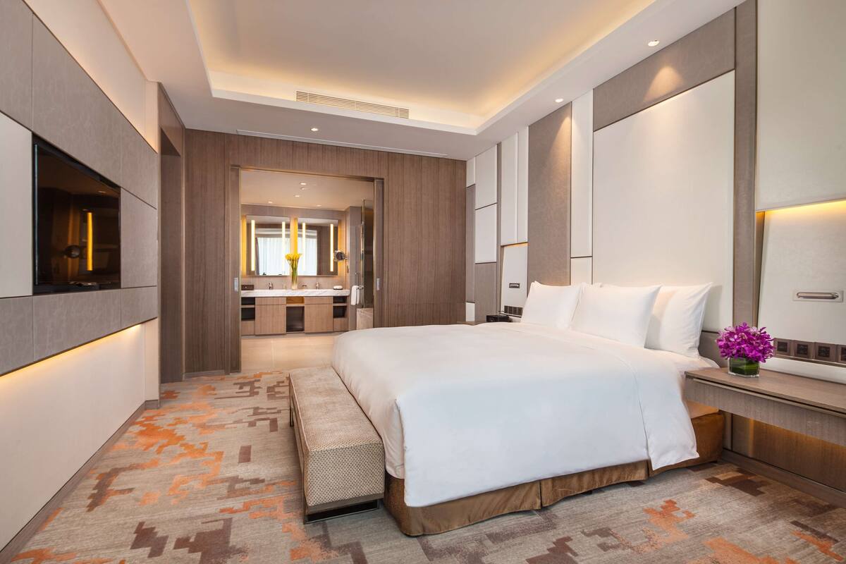 A modern hotel room with a large bed, a bench, a vanity with a mirror, and a television.
