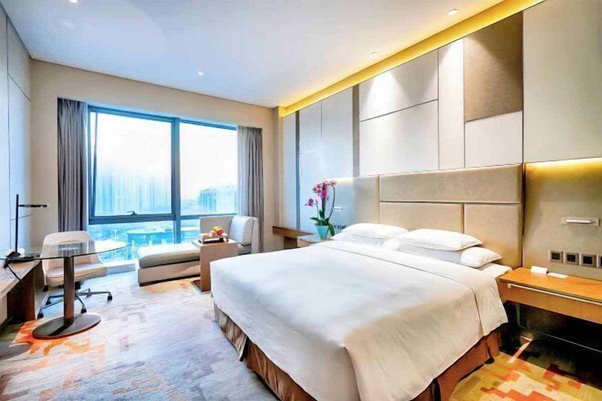 A modern hotel room with a large bed, a desk, and a view of a cityscape.