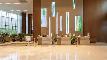 A hotel reception area with a marble floor, reception desk, potted plants, and large windows with vertical blinds.