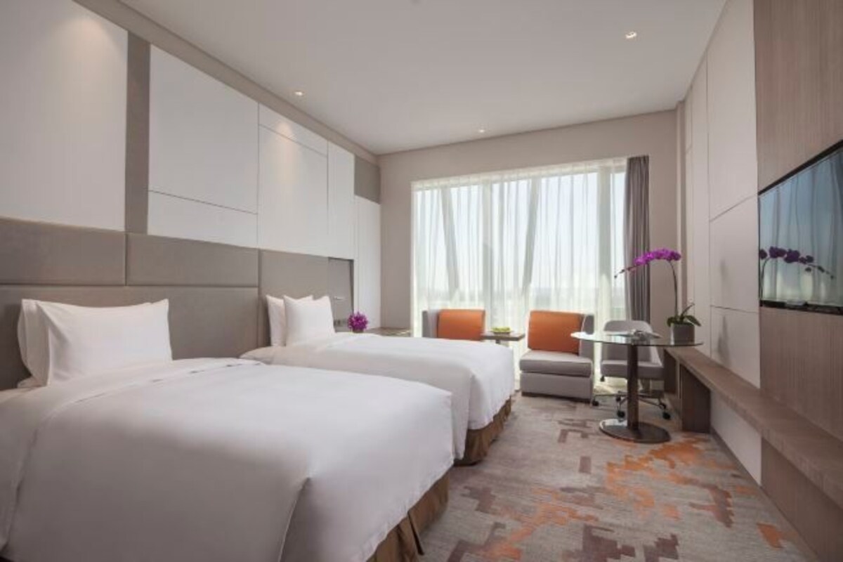 Habitación de hotel con dos camas, una mesita, una silla y un ventanal grande con cortinas.