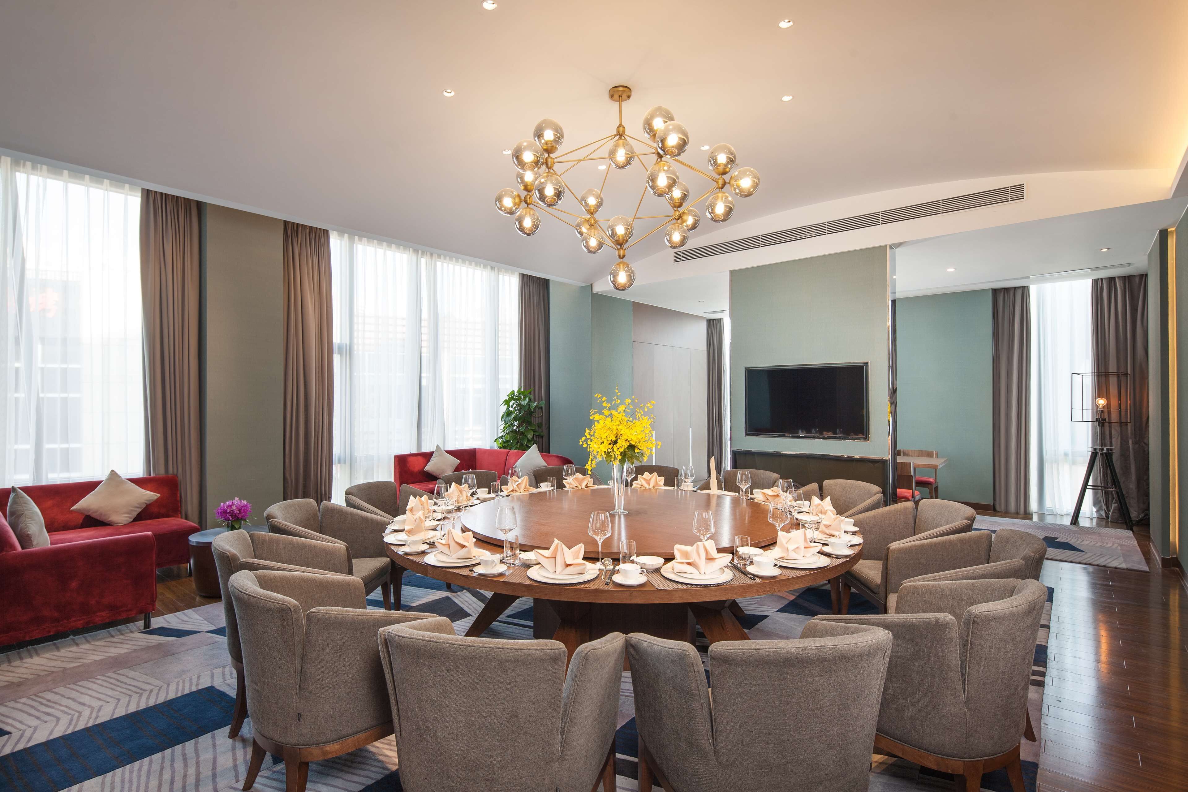 Amplia habitación de hotel con una mesa redonda de comedor preparada para seis personas, rodeada de seis sillas. Hay un sofá rojo y un televisor en la habitación.