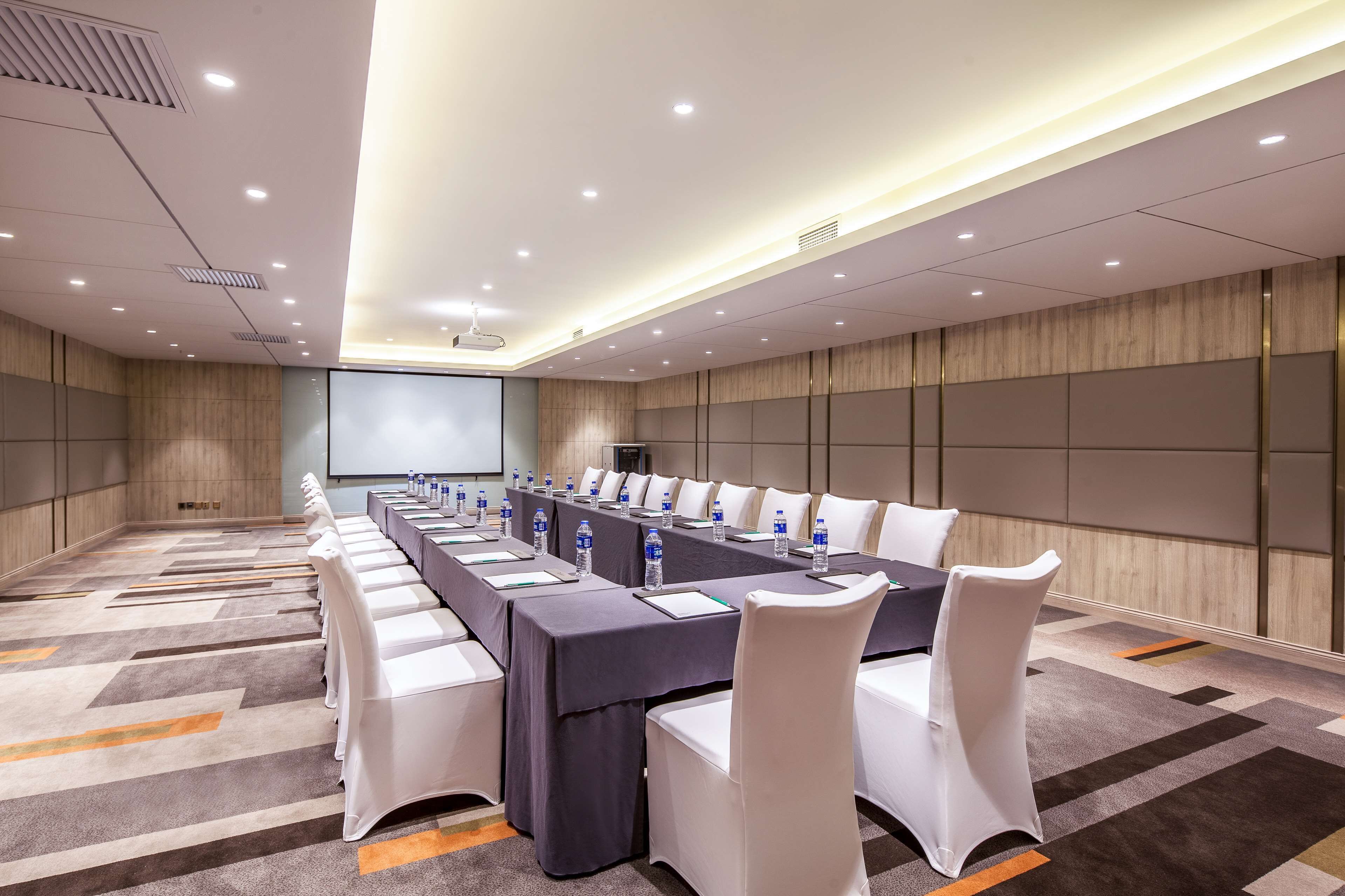 Uma sala de conferências com uma mesa comprida, cadeiras brancas e uma tela de projeção.