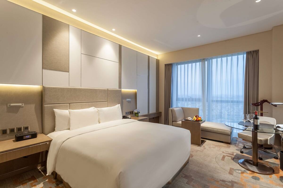 A hotel room with a large bed, a desk with a chair, a small table with a lamp, and a large window with curtains.
