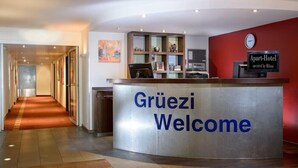 A hotel reception area with a curved reception desk labeled "Grüezi Welcome." - Apart Hotel Zurich Airport (Opfikon)
