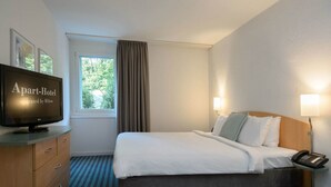 A hotel room with a bed, a television, a desk with drawers, a telephone, and a window with curtains. - Apart Hotel Zurich Airport (Opfikon)