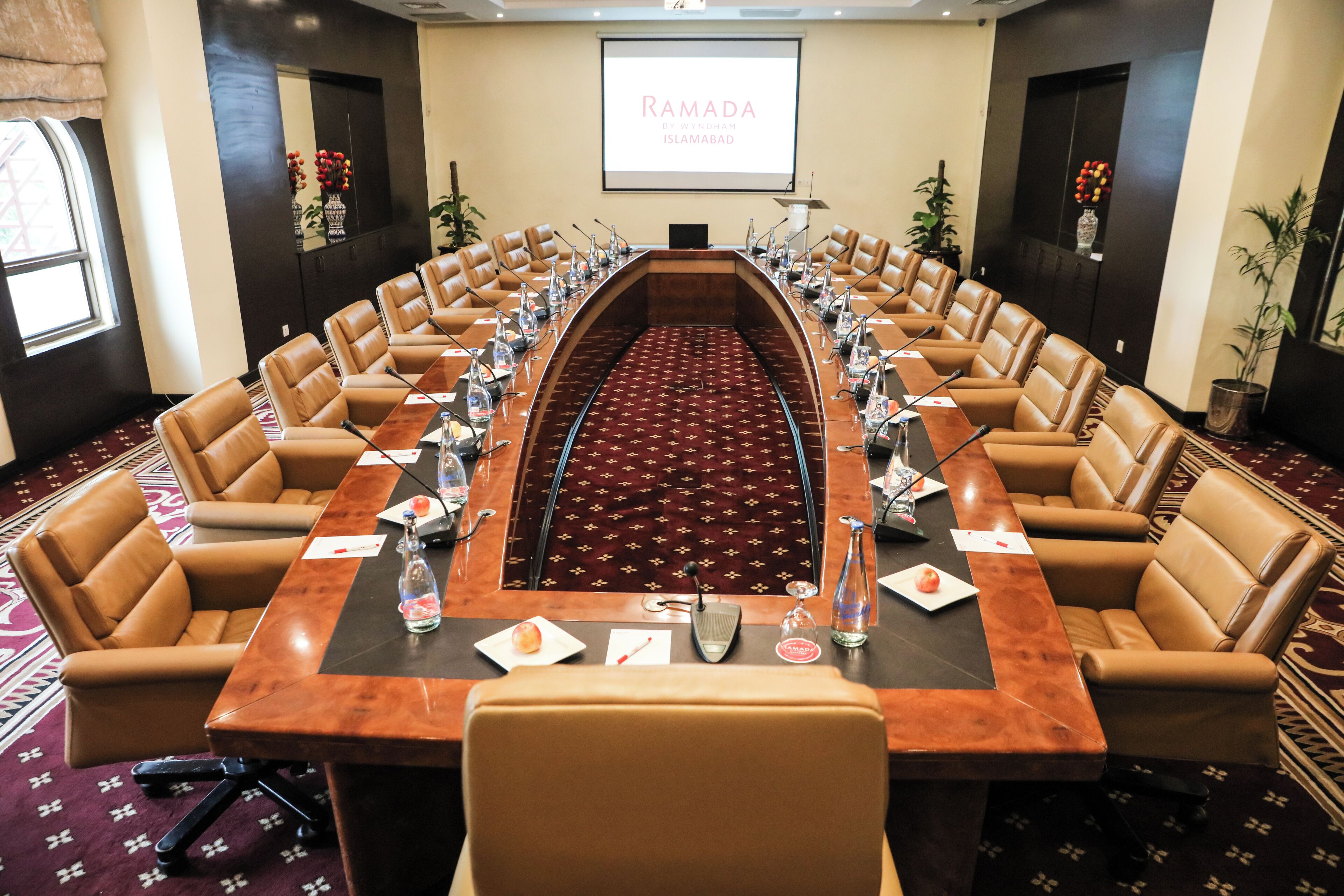 A conference room with a long table, leather chairs, and a projector screen displaying "RAMADA".