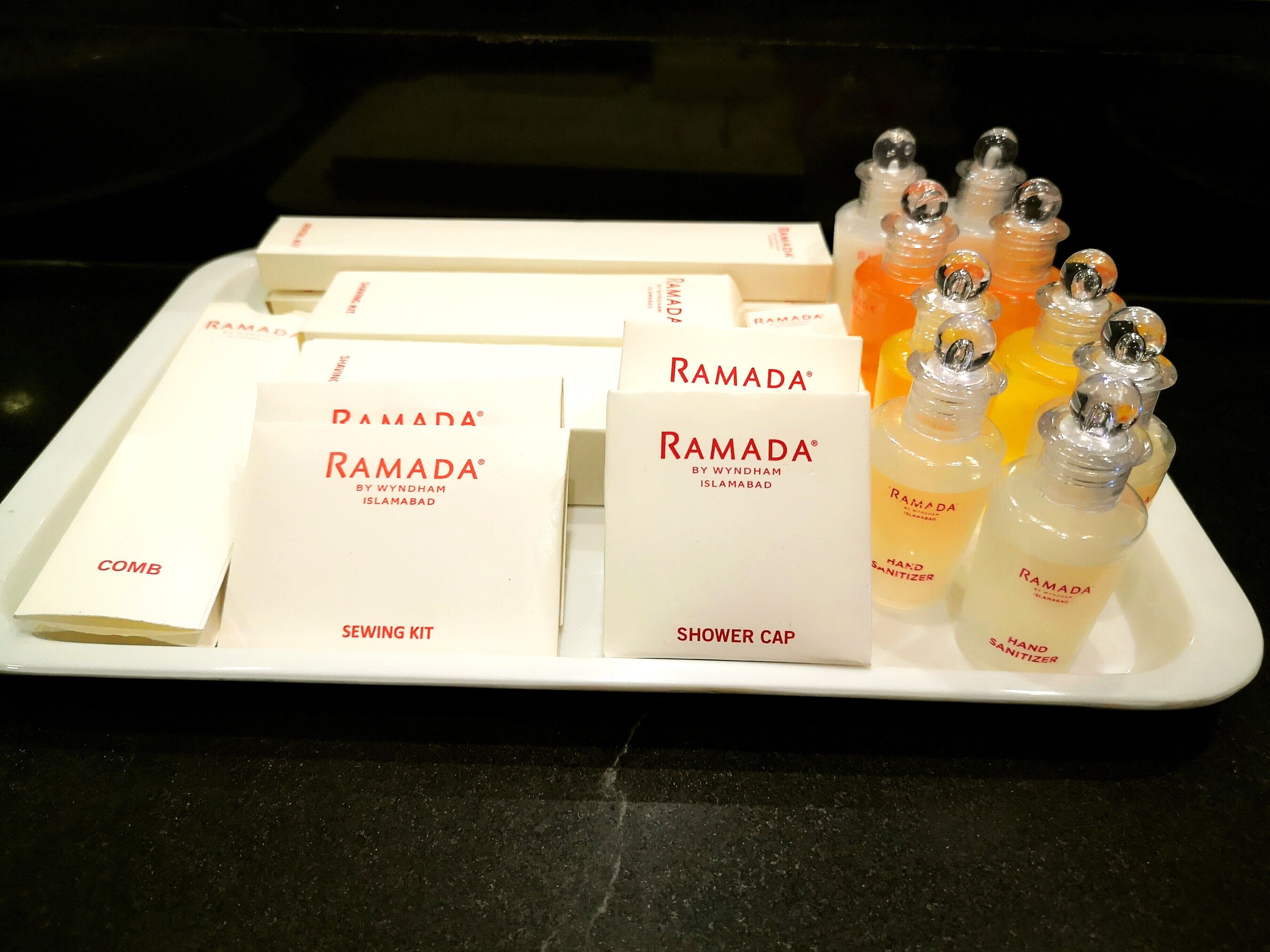 A tray with various Ramada® branded items including a sewing kit, a shower cap, and hand sanitizer bottles.