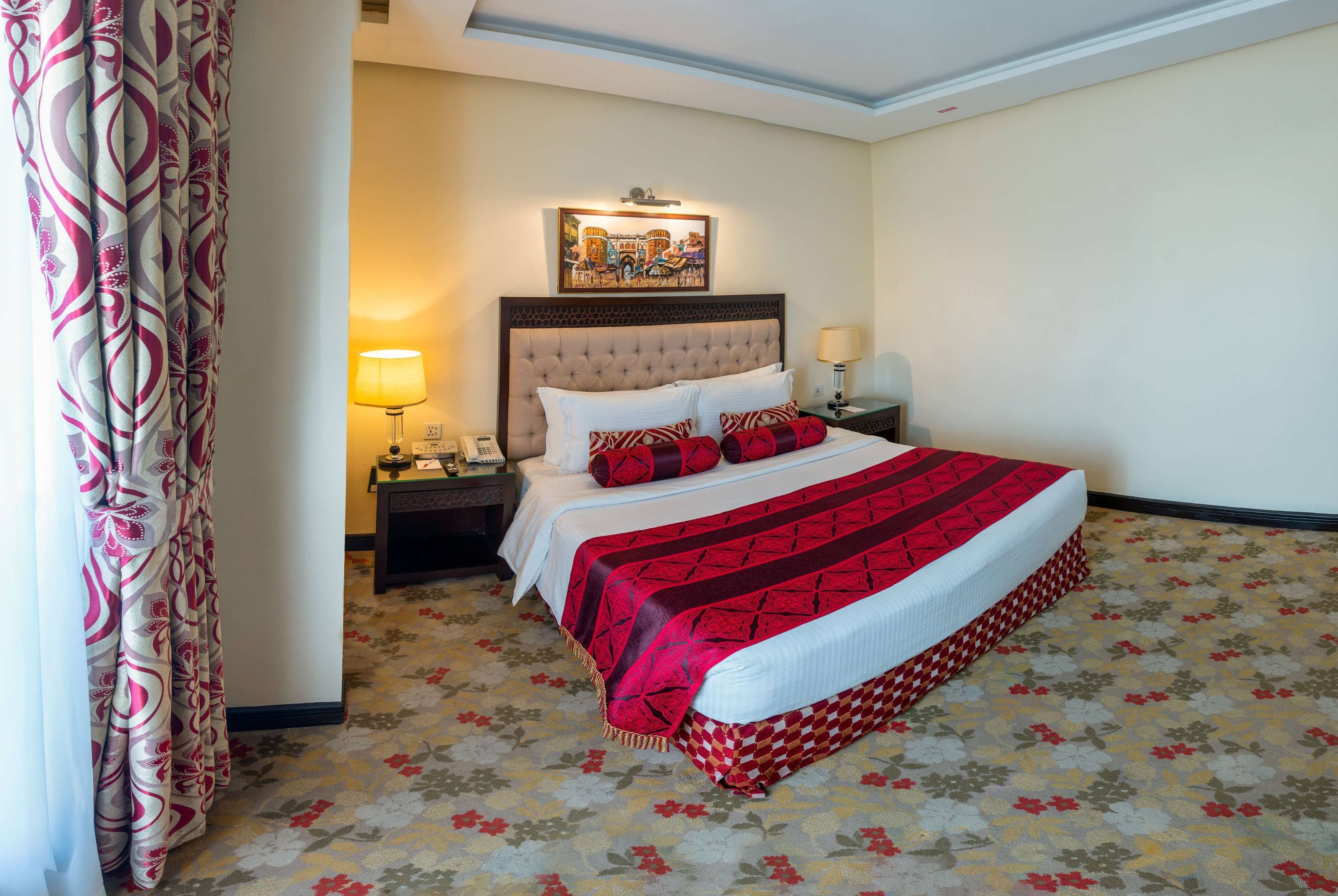 Hotelkamer met een groot bed, nachtkastje met lamp, ingelijste foto aan de muur en een bloemenprint tapijt.